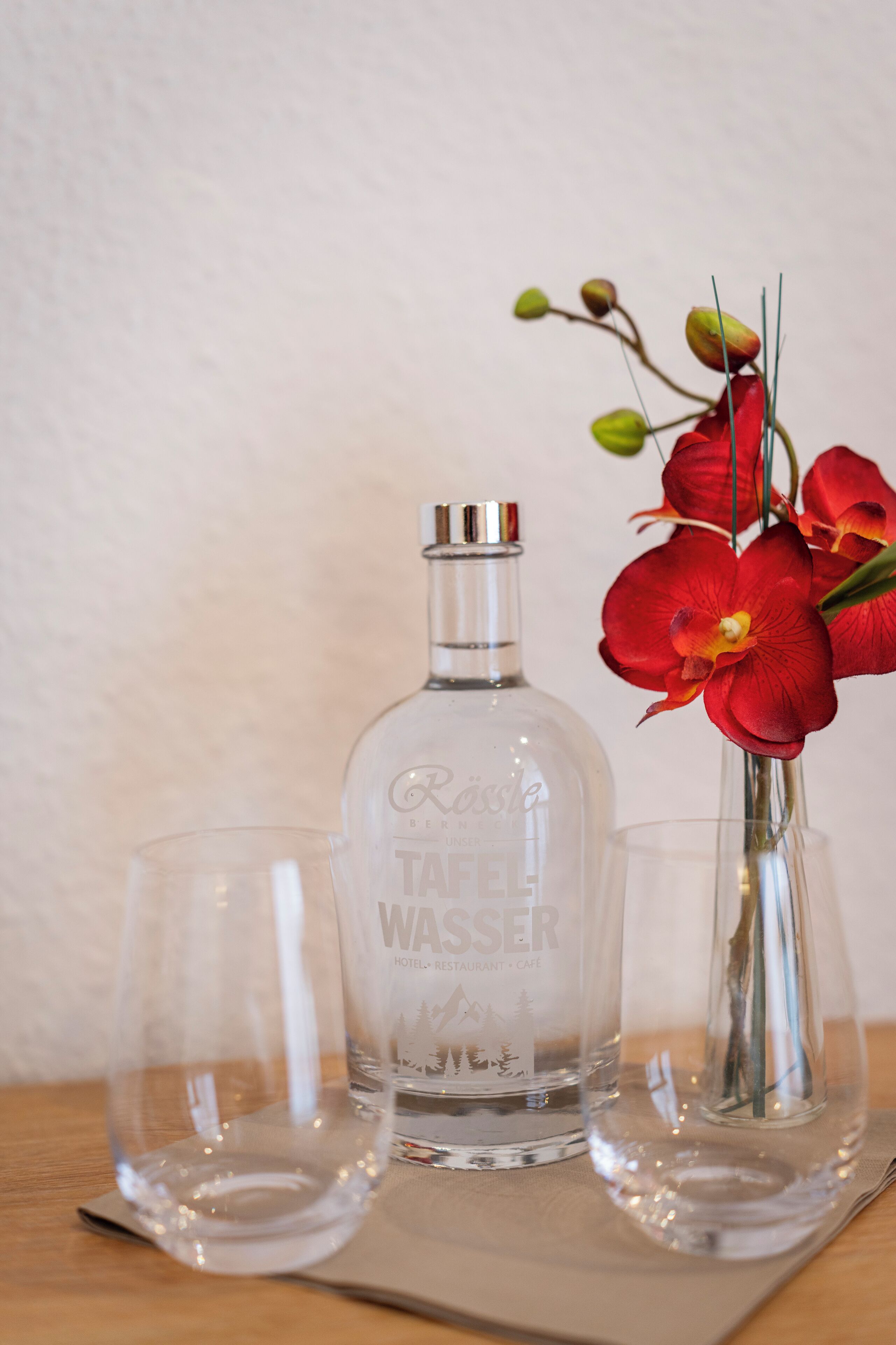 A bottle of water, two glasses, and a vase with red flowers on a wooden table.