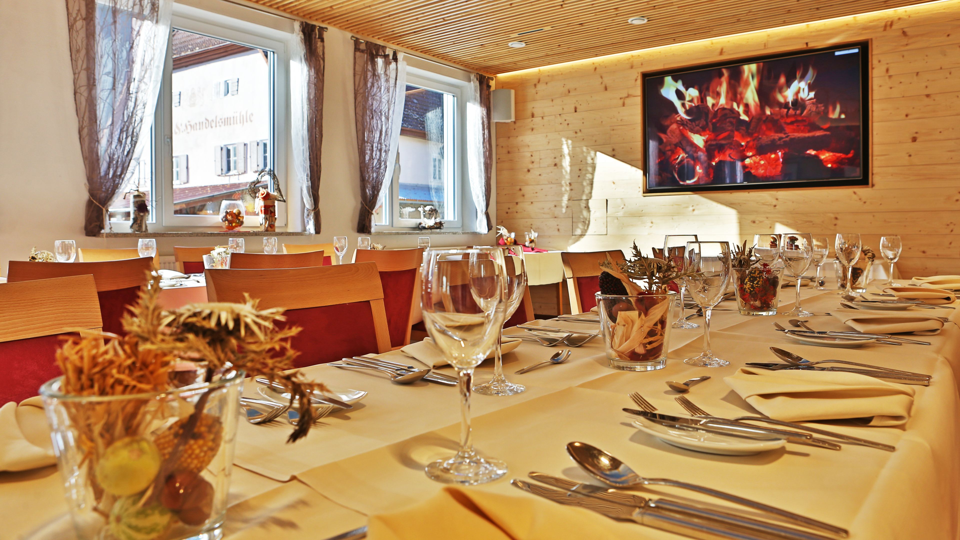Ein Restaurant mit Holzwänden und einem Fernseher, der ein Feuer zeigt. Die Tische sind mit Weingläsern, Besteck und Servietten gedeckt.