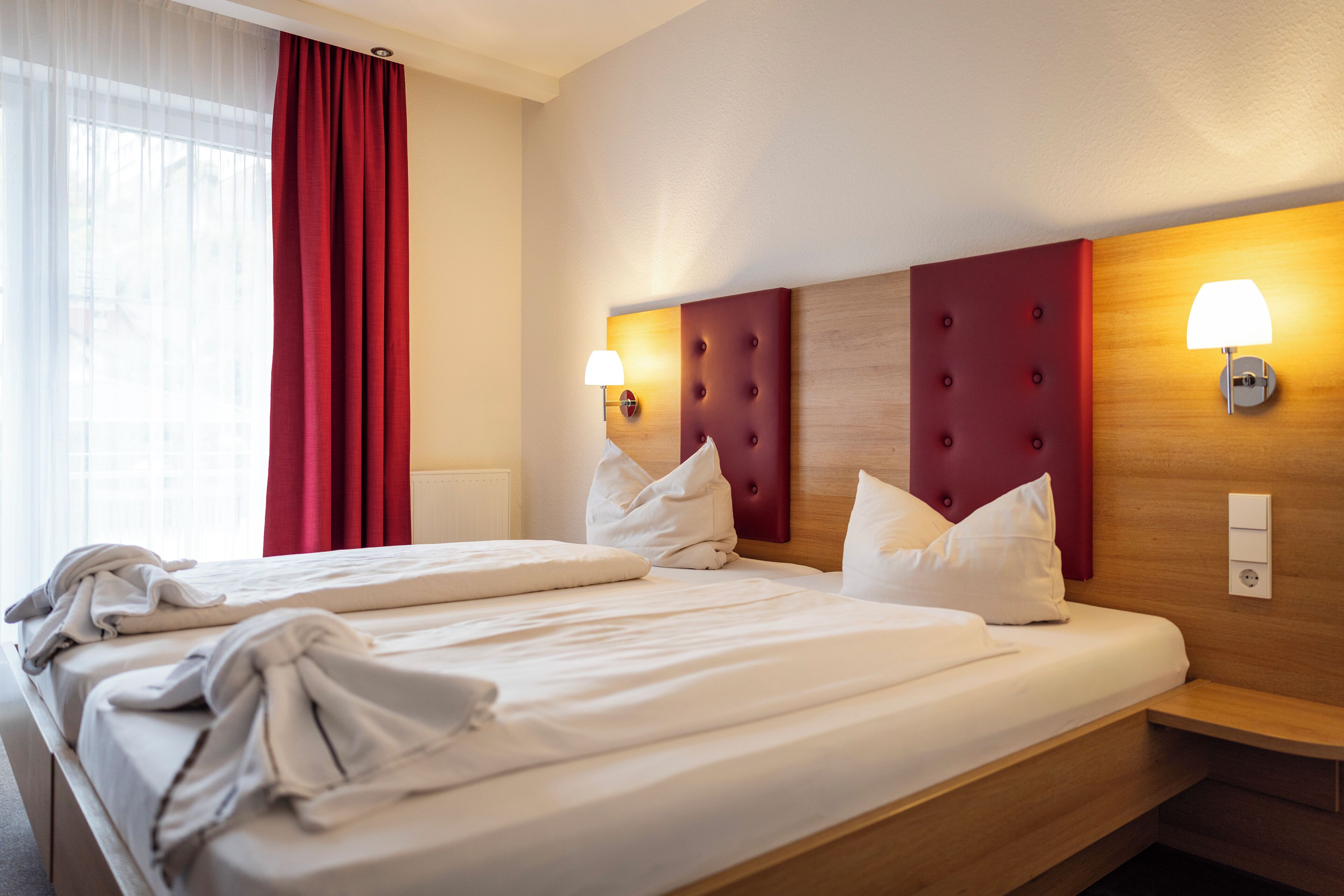 A hotel room with two beds, a wooden headboard, bedside lamps, and a red curtain.
