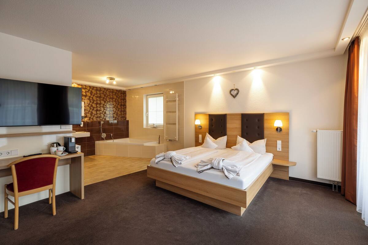 A modern hotel room with a large bed, a desk, a chair, a TV, and a bathroom.