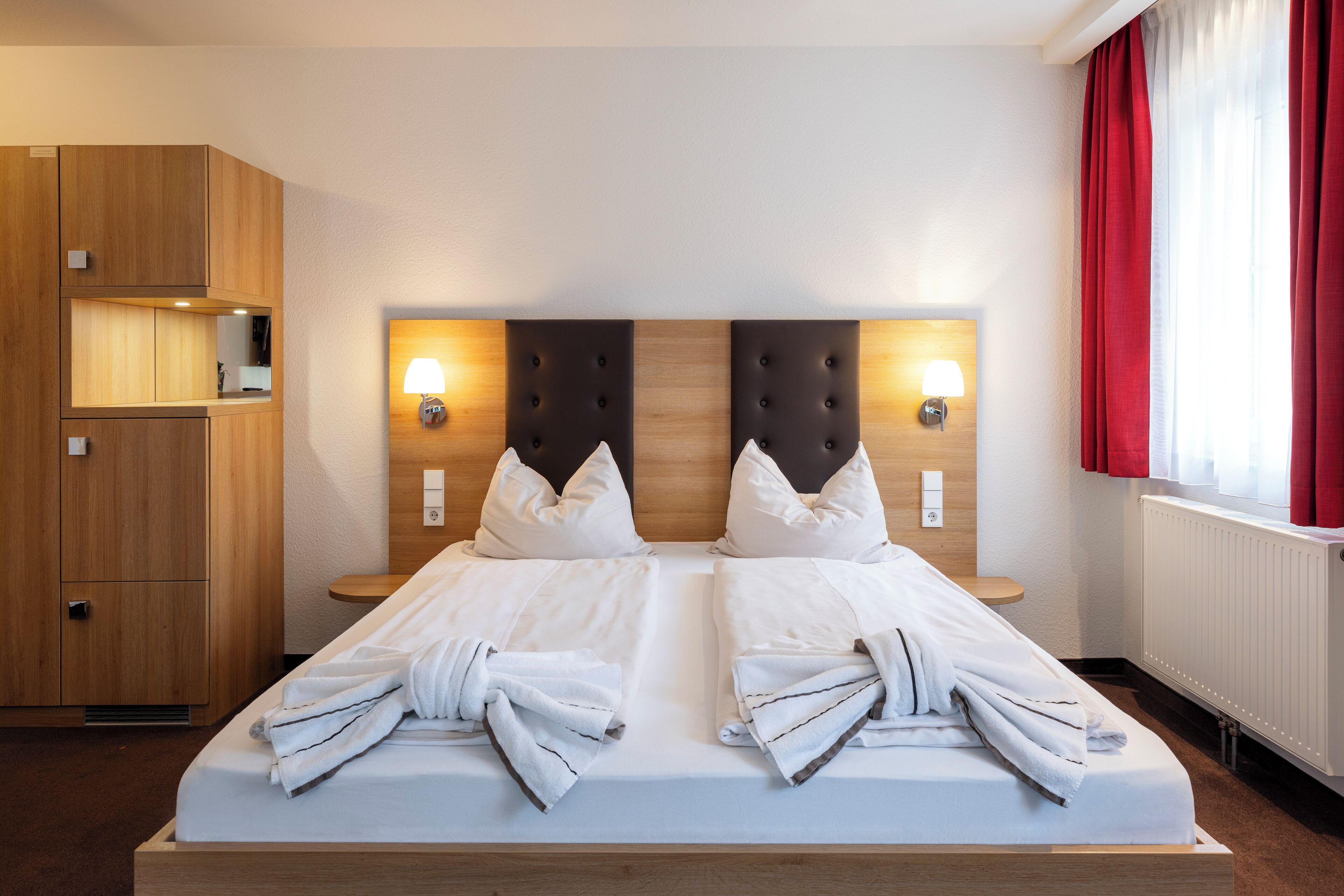 Een tweepersoonsbed met witte lakens en handdoeken, een houten hoofdbord, nachtlampjes en een rood gordijn.
