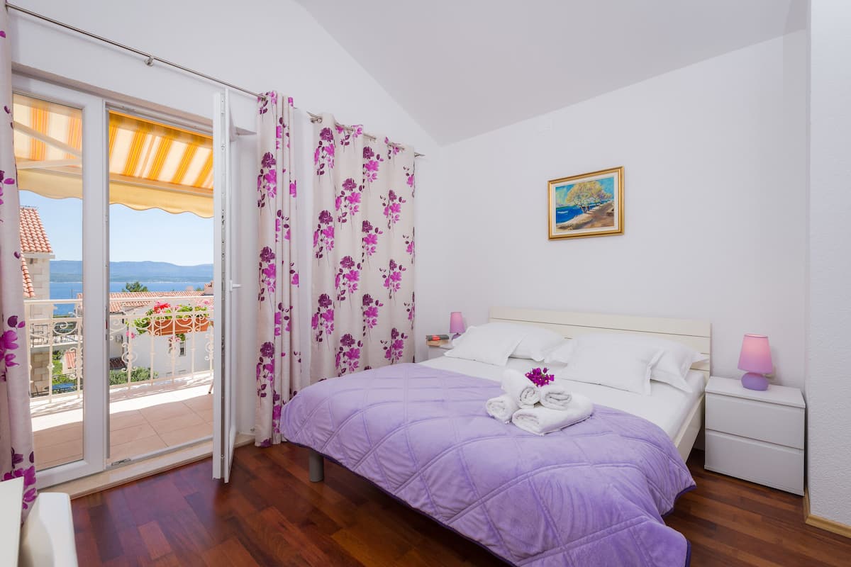 Un dormitorio con cama, mesita de noche, lámpara, un cuadro y vista a un balcón con barandilla y un edificio.