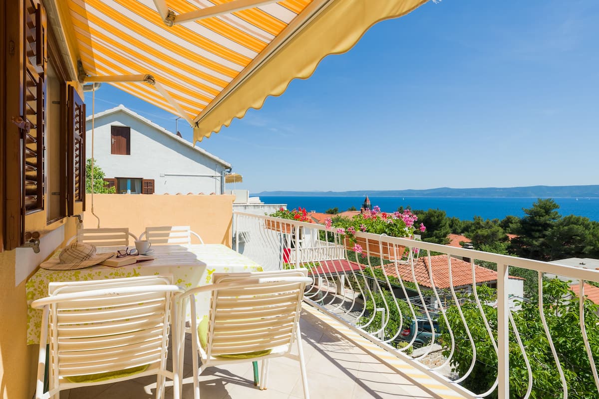 Un balcón con mesa y sillas, con vistas a la costa, casas y un cielo despejado.