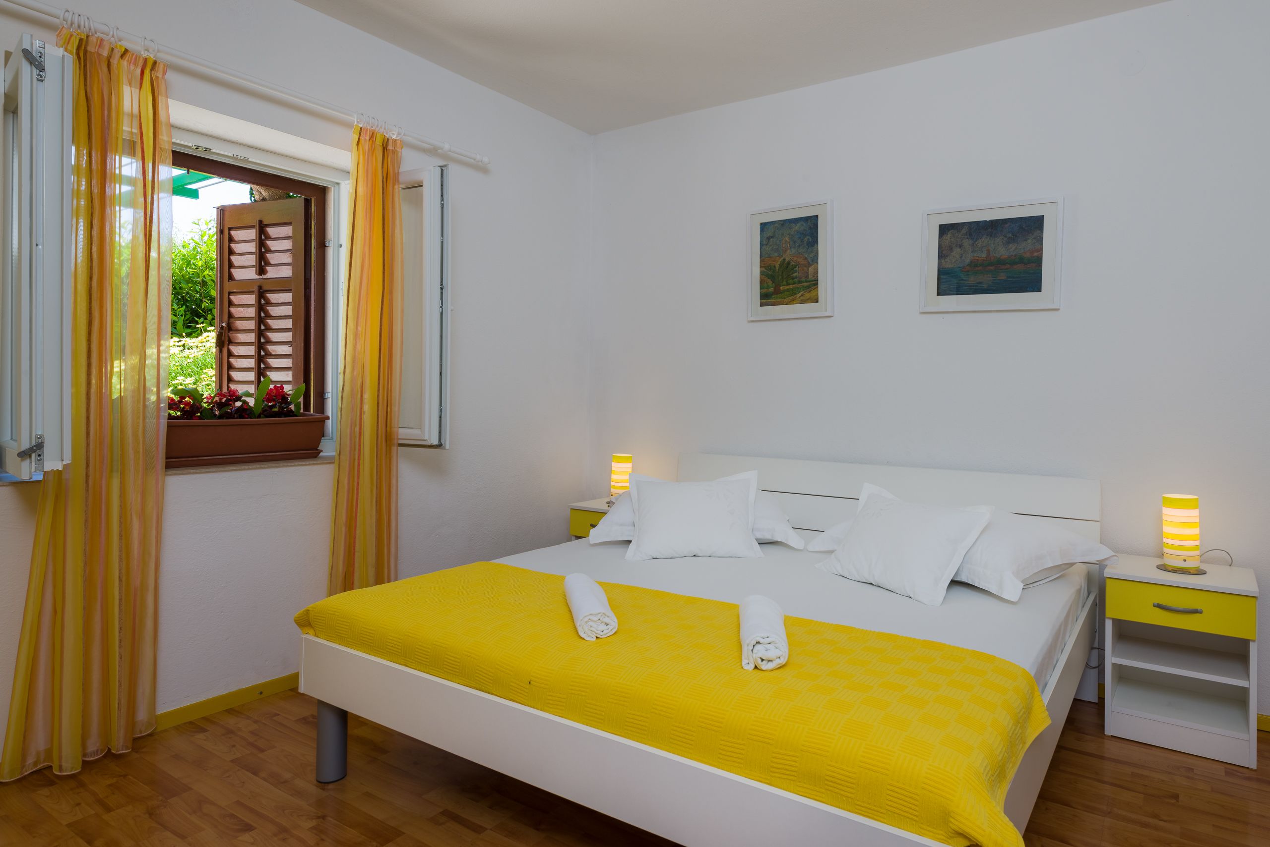 A neatly made bed with white linens and a yellow bedspread, a window with yellow curtains, and two bedside lamps.