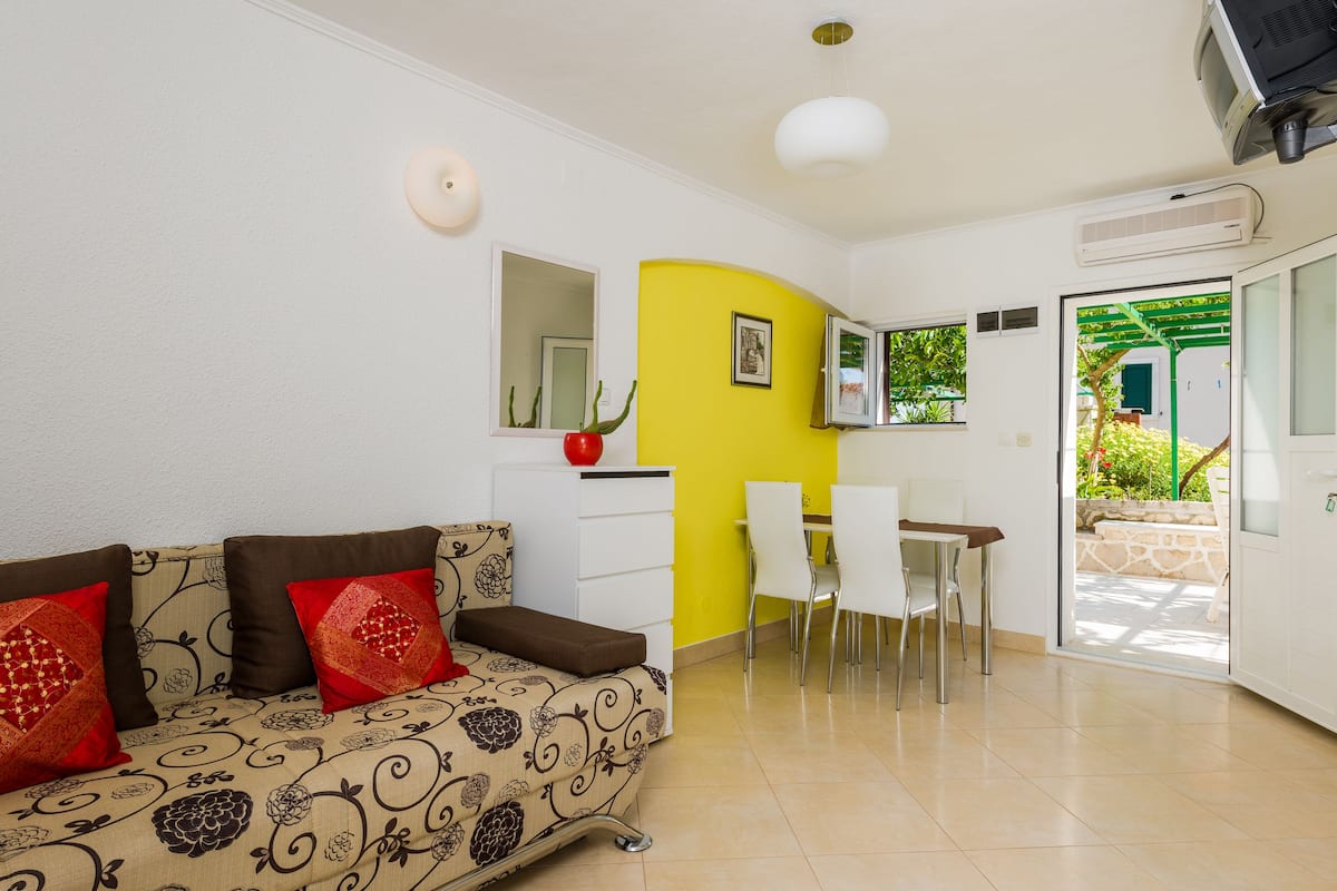Una sala de estar con sofá, mesa de comedor y sillas. Se ve vegetación por la ventana.