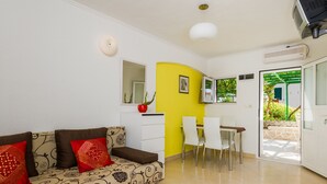 Una sala de estar con sofá, mesa de comedor y sillas. Se ve vegetación por la ventana.