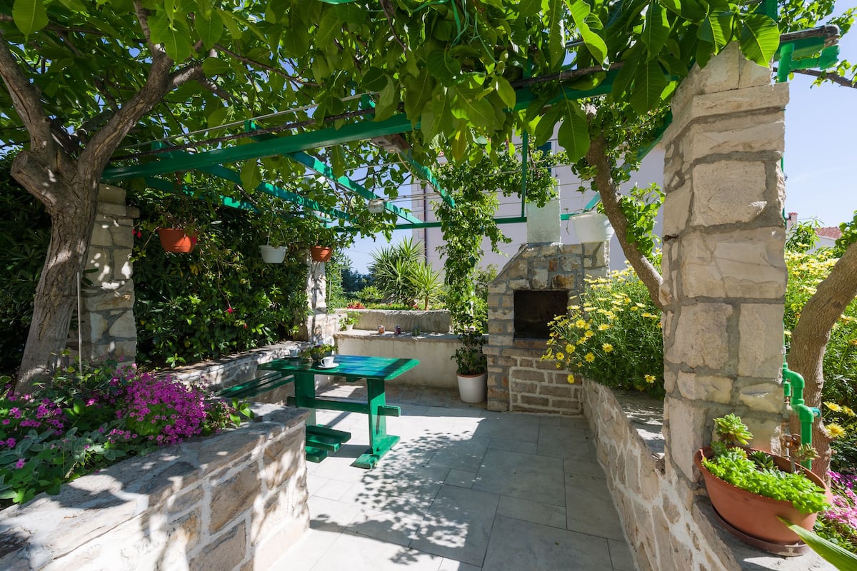 Un patio de piedra con una mesa y sillas verdes, rodeado de macetas con plantas y un muro de piedra.