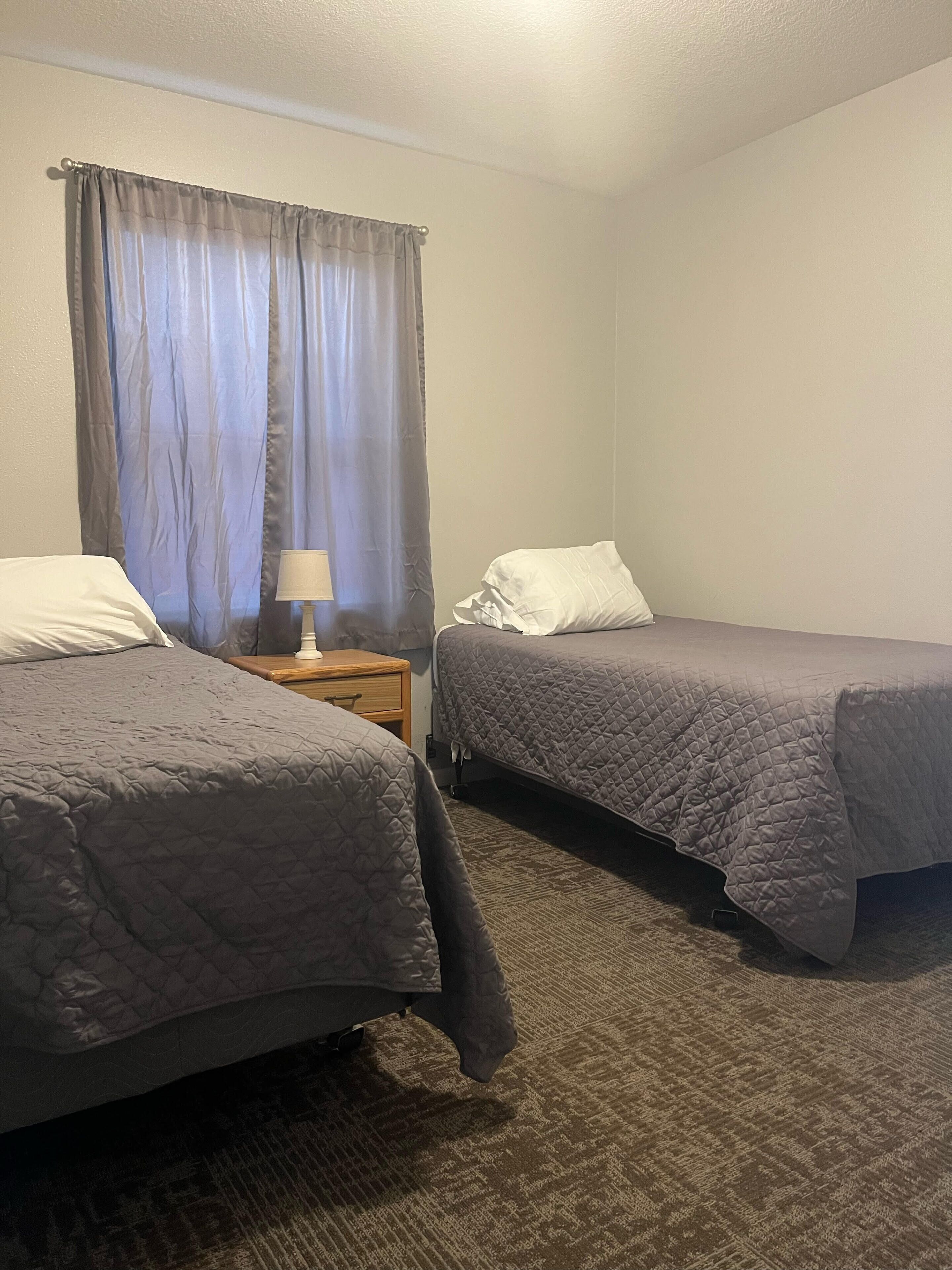 A bedroom with two single beds, a nightstand with a lamp, and a window with curtains.