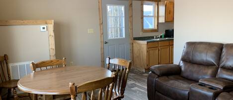 A dining area with a wooden table and chairs, a brown leather sofa, and a kitchen with wooden cabinets and a window.