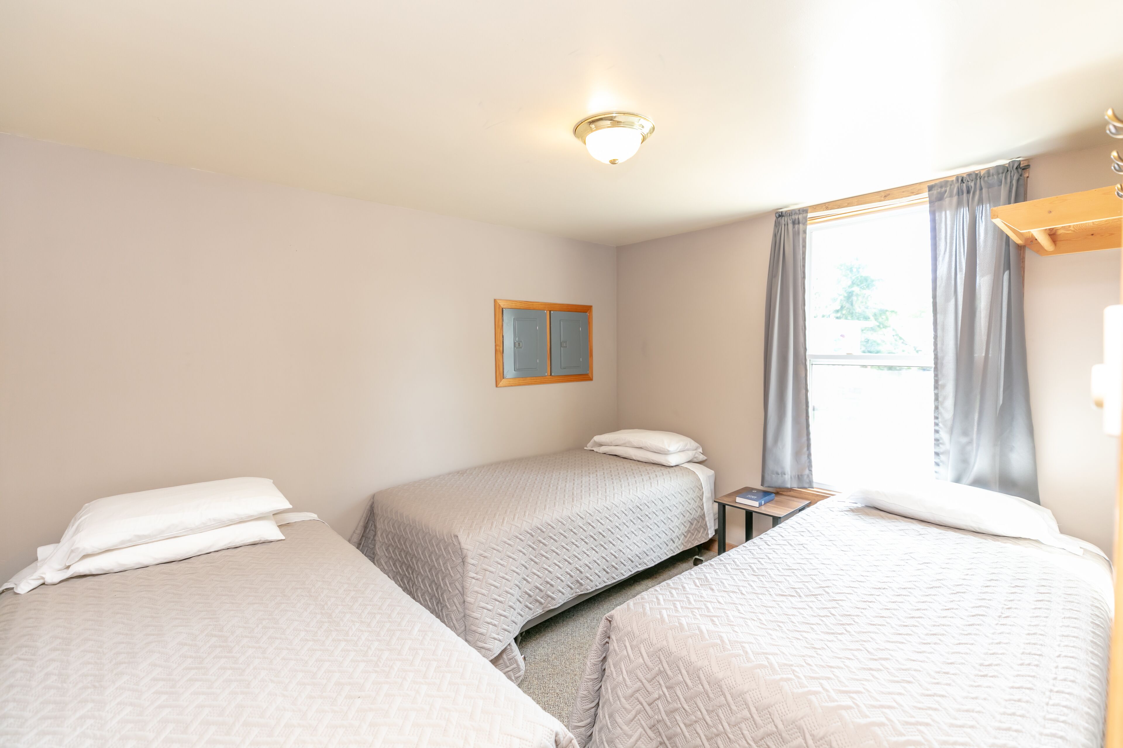 A hotel room with two beds, a small table, a nightstand, a window with curtains, and a ceiling light fixture.