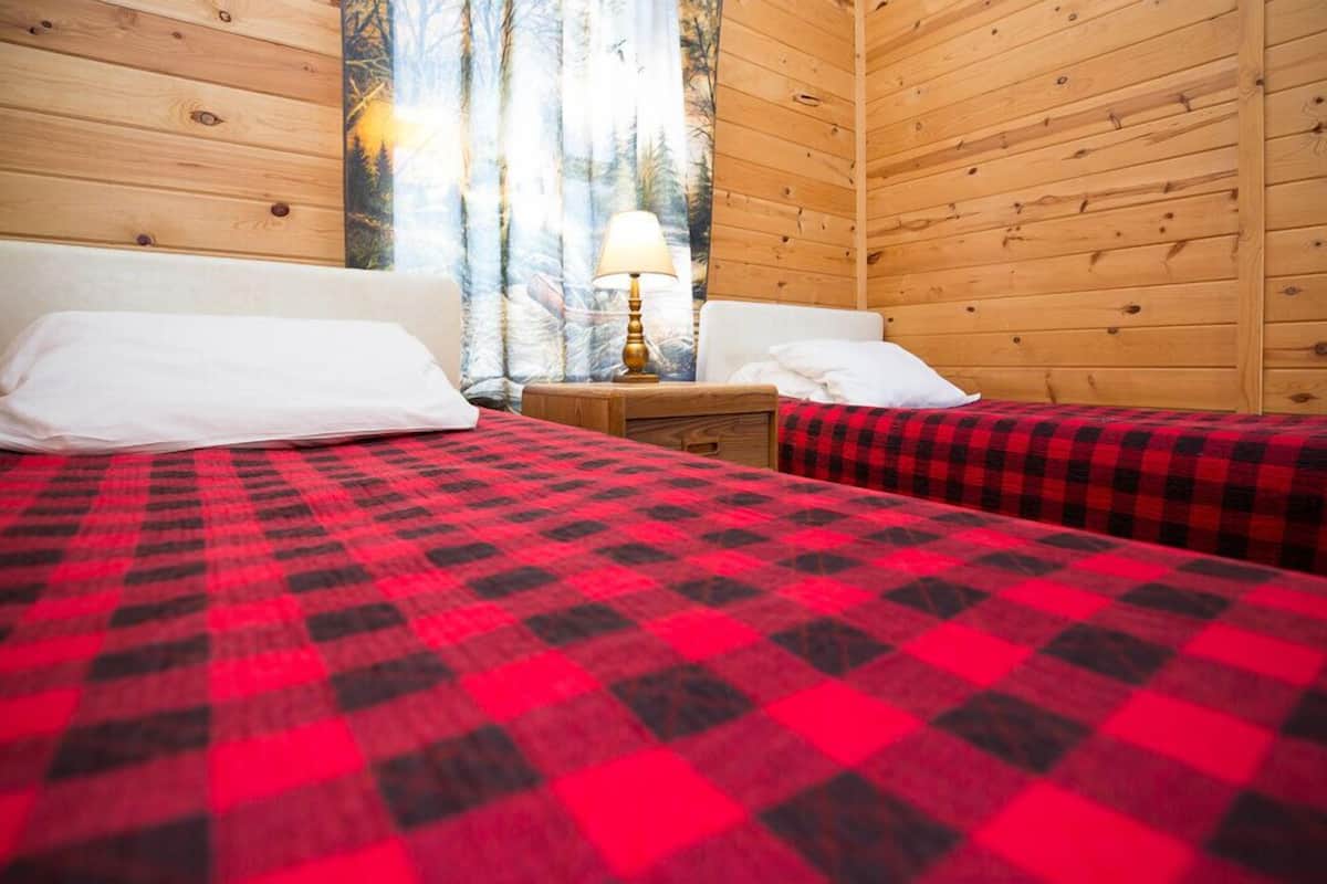 Quarto estilo cabana com duas camas, cada uma com colcha xadrez vermelha e preta, paredes de madeira, mesa de cabeceira com abajur e uma janela com cortinas.