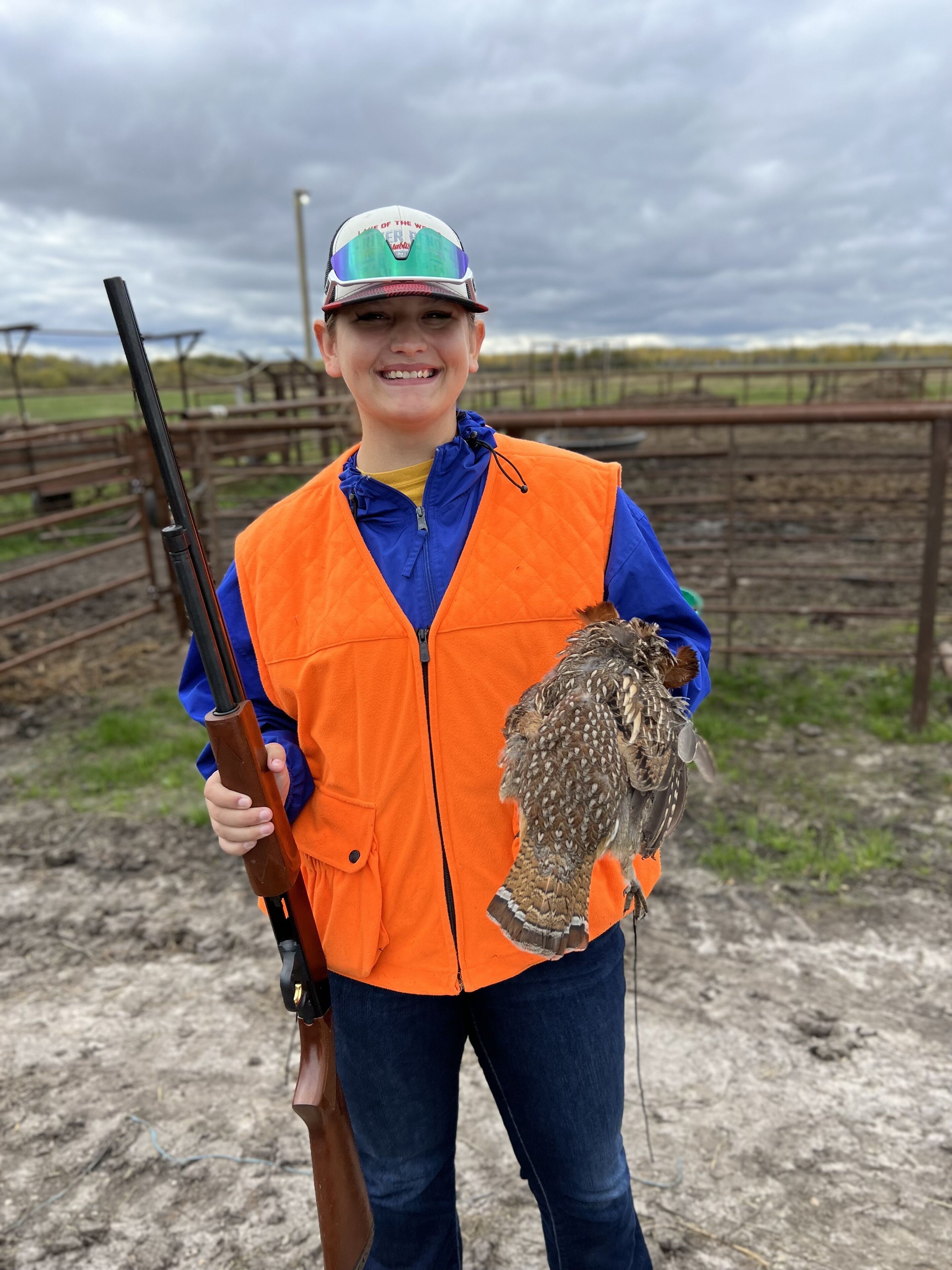 A person holding a rifle and an owl, standing in an outdoor area with a fence and grassy field in the background.