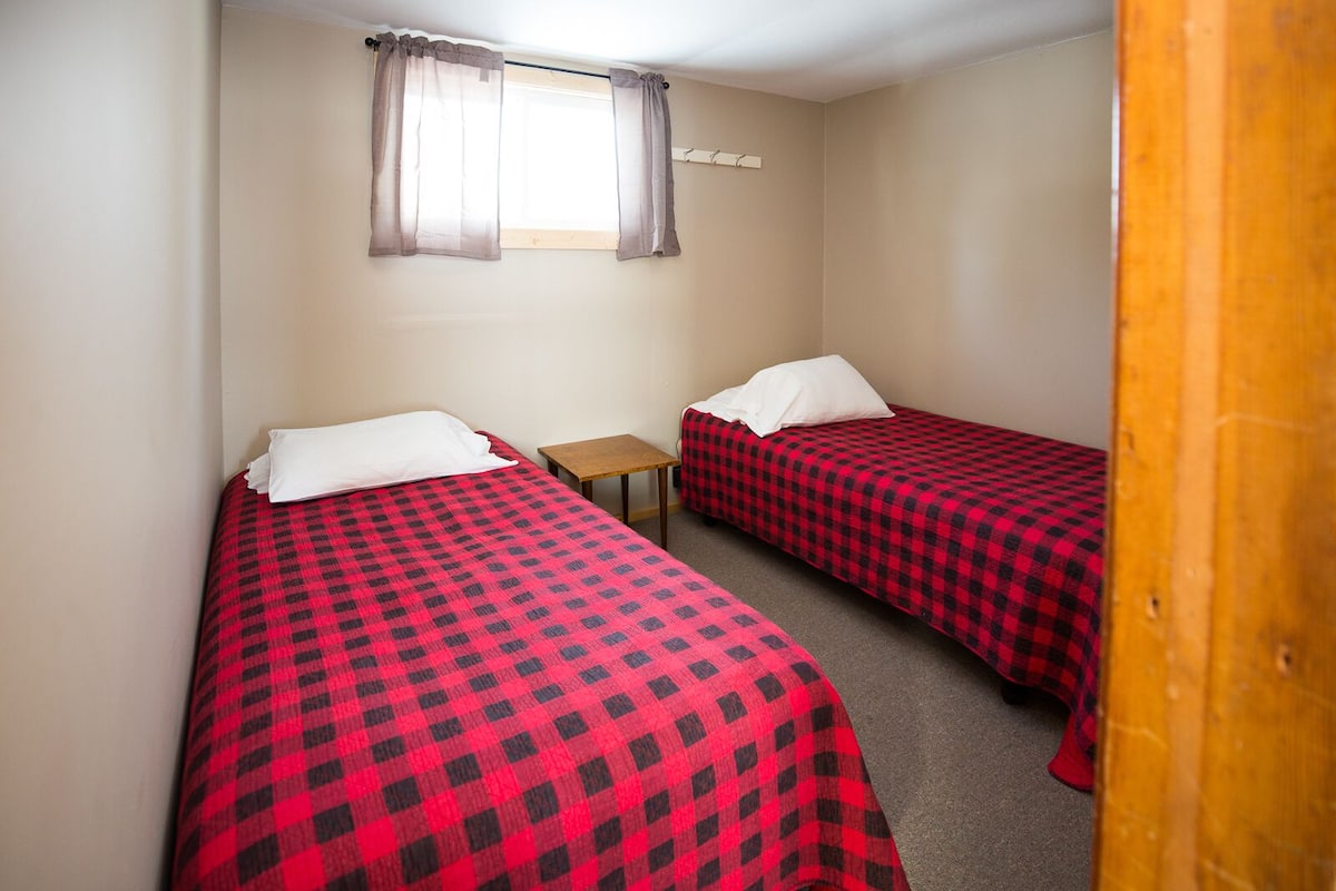 Quarto pequeno com duas camas de solteiro, cada uma com colcha xadrez vermelha e preta e almofadas brancas. Há uma pequena mesa de madeira entre as camas, e uma janela com cortinas leves acima.