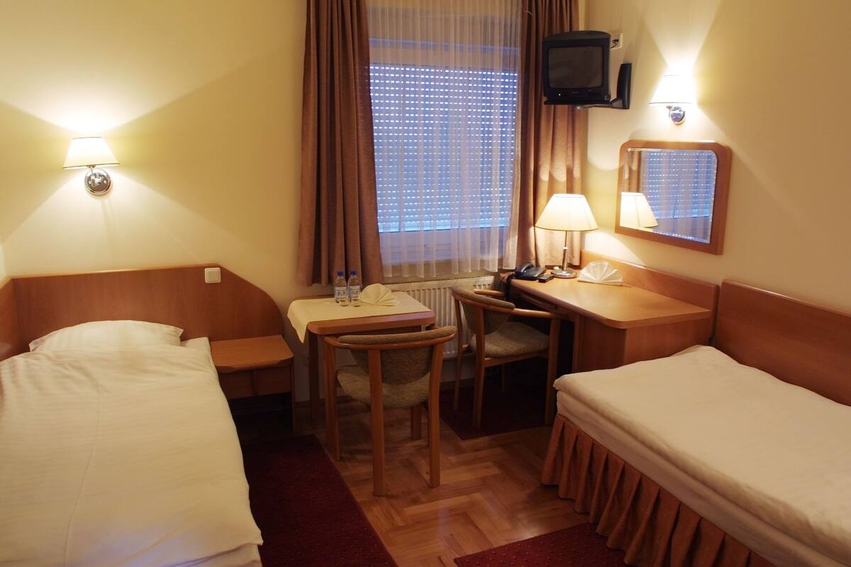 A hotel room with two single beds, a desk with a chair, a small table, a telephone, and a television mounted on the wall.