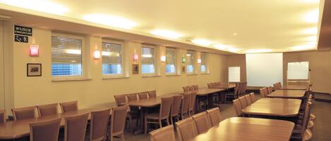A conference room with multiple tables and chairs, equipped with ceiling lights and windows with blinds.