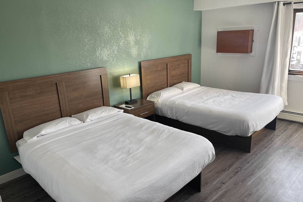 A hotel room with two beds, a nightstand with a lamp, and a view of the outdoors through a window with curtains.