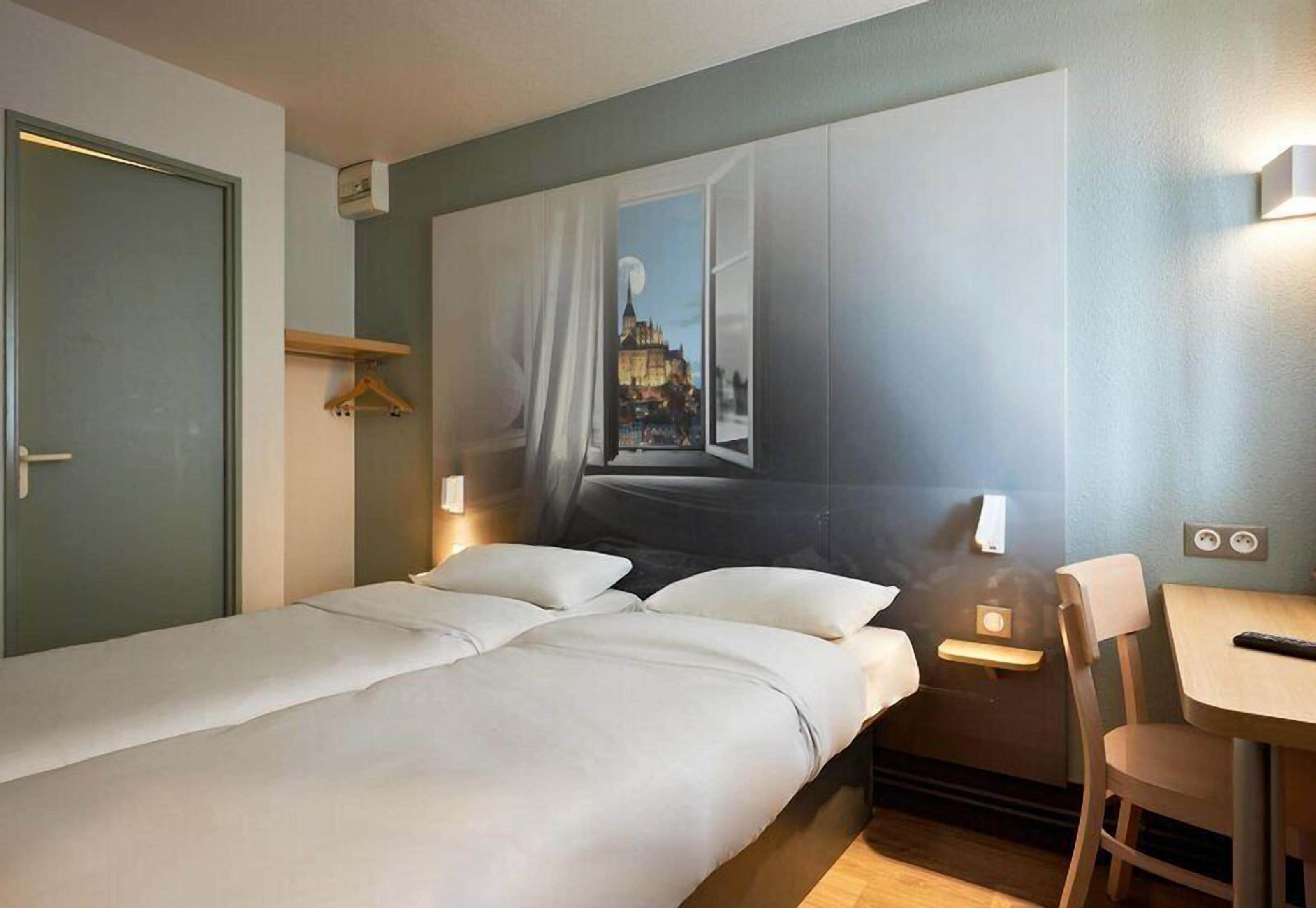 A hotel room with a bed, bedside tables, a chair, a desk, a painting on the wall, and a door.