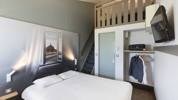 A hotel room with a bed, a TV mounted on the wall, a coat rack with clothes, and a staircase leading to an upper level.