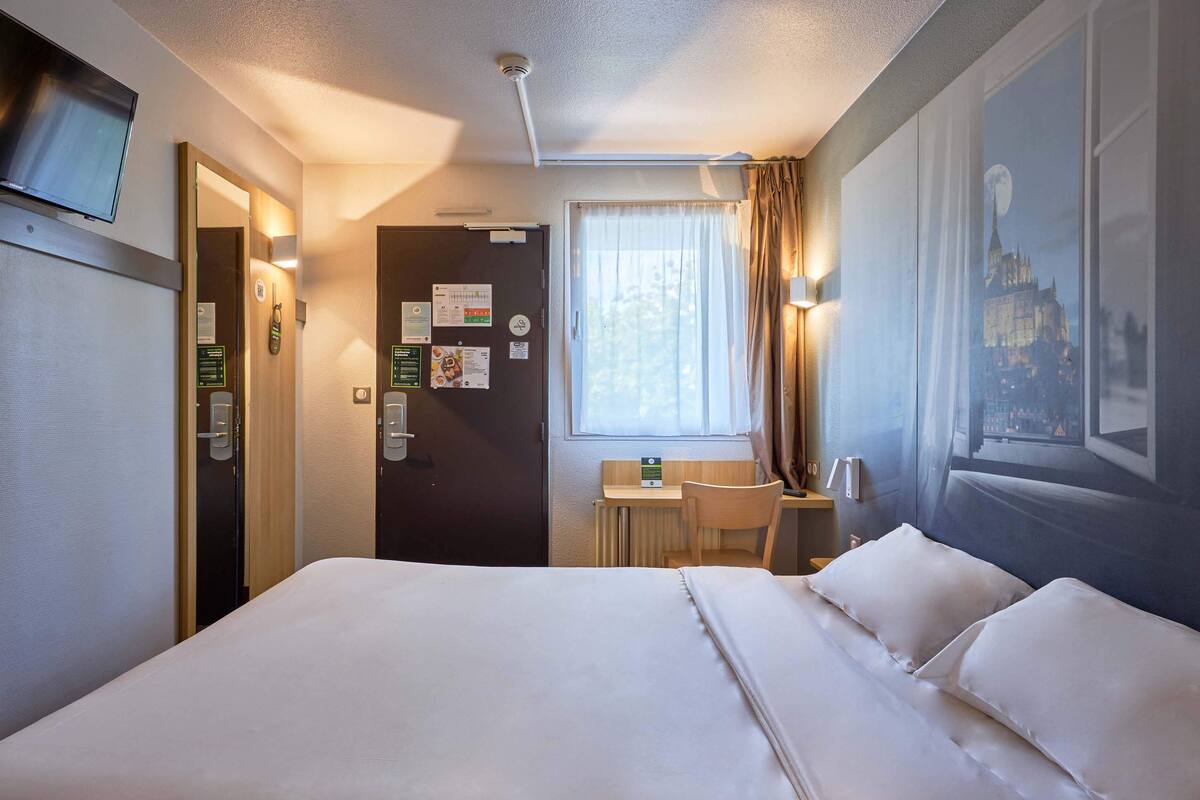 Una habitación de hotel con una cama grande, un escritorio con silla, una ventana con cortinas y un televisor montado en la pared.
