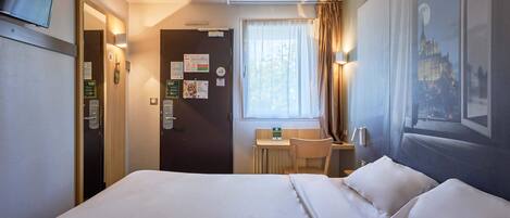 A hotel room with a large bed, a desk with a chair, a window with curtains, and a wall-mounted TV.