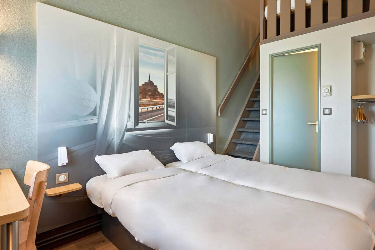 Una habitación de hotel con una cama grande, un escritorio, una silla, una mesita de noche, una lámpara de pared y una escalera.