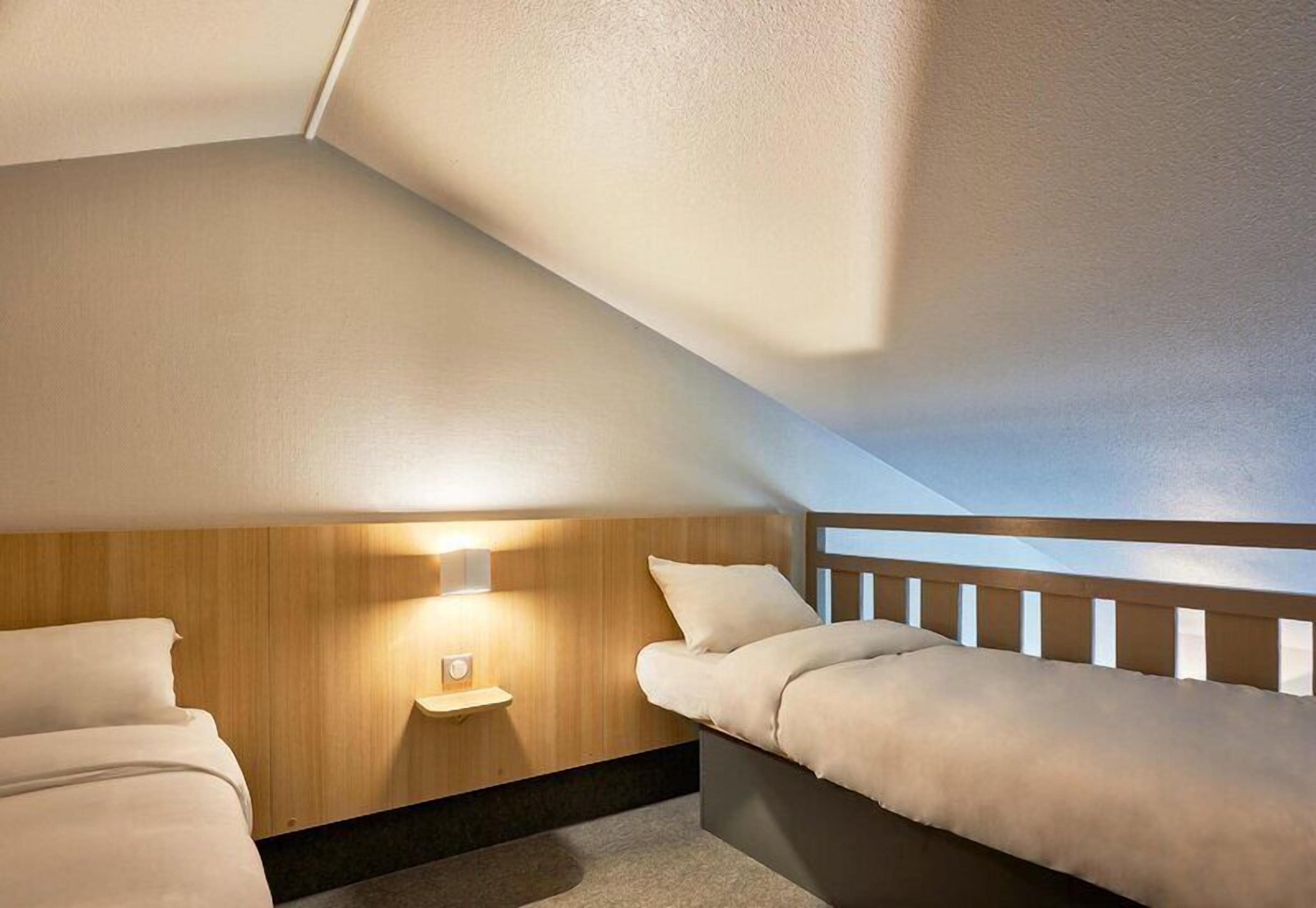 A small, cozy room with a sloped ceiling, a wooden headboard, a bedside table with a lamp, and a cushioned bench.
