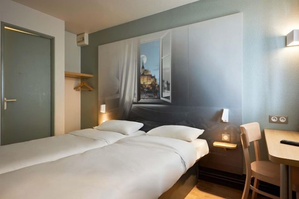Habitación de hotel con cama, escritorio y silla. Se ve una ventana con vistas a un paisaje urbano.