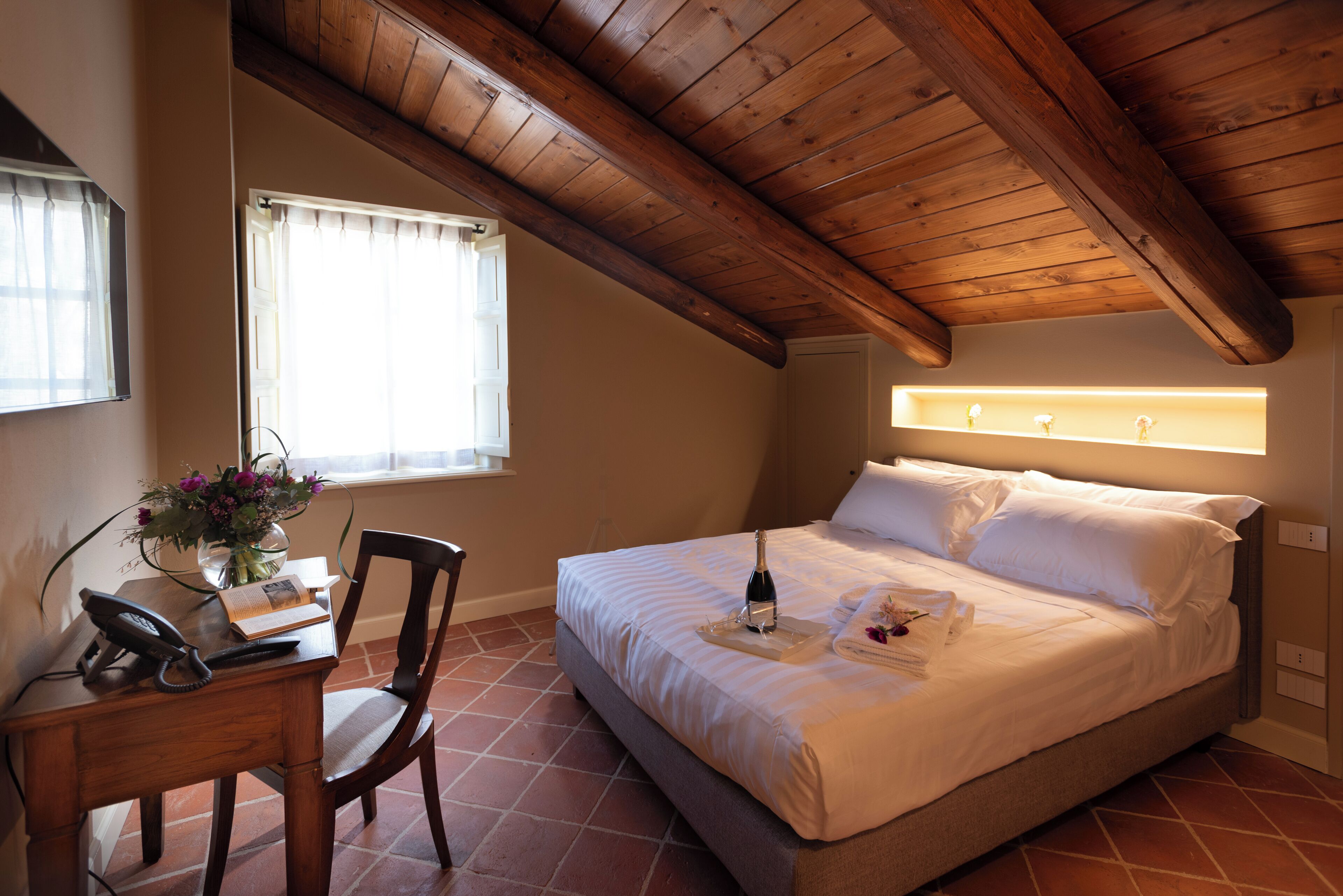 Een slaapkamer met een houten plafond, een bed met witte lakens, een nachtkastje met telefoon en boek, en een raam met gordijnen.