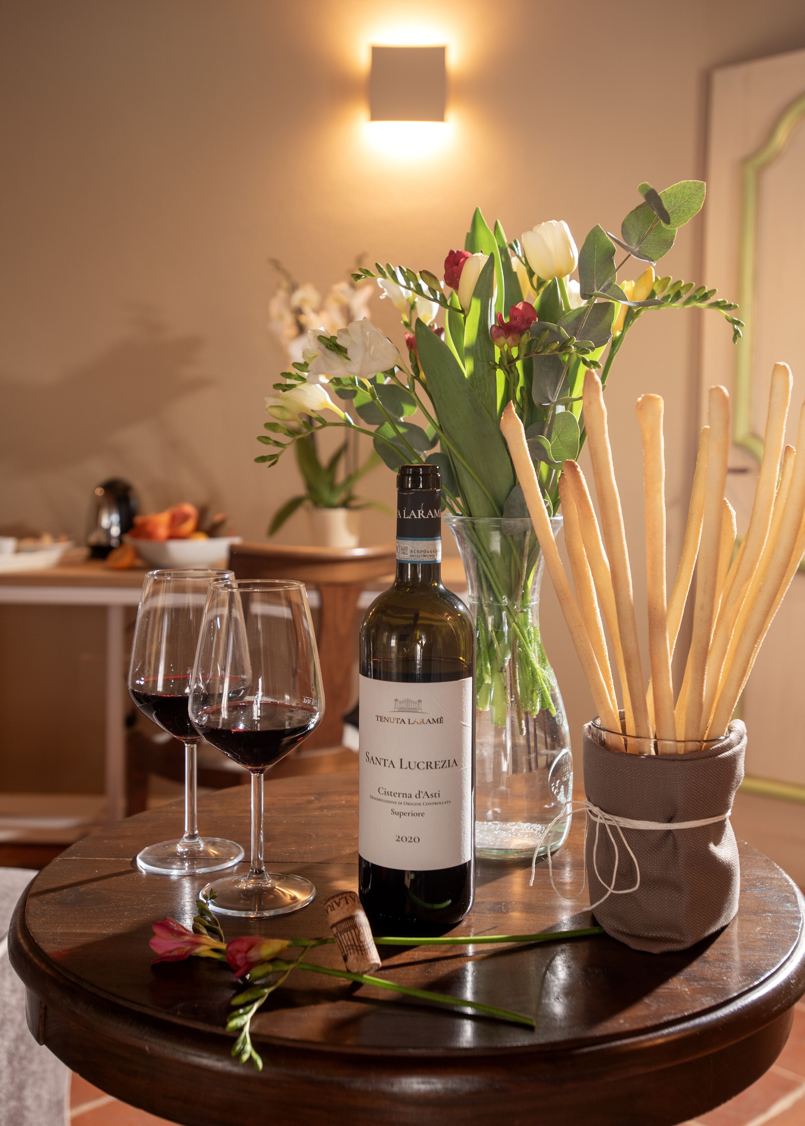 A table with a bottle of wine, two glasses of red wine, a vase with flowers, and a container of breadsticks.