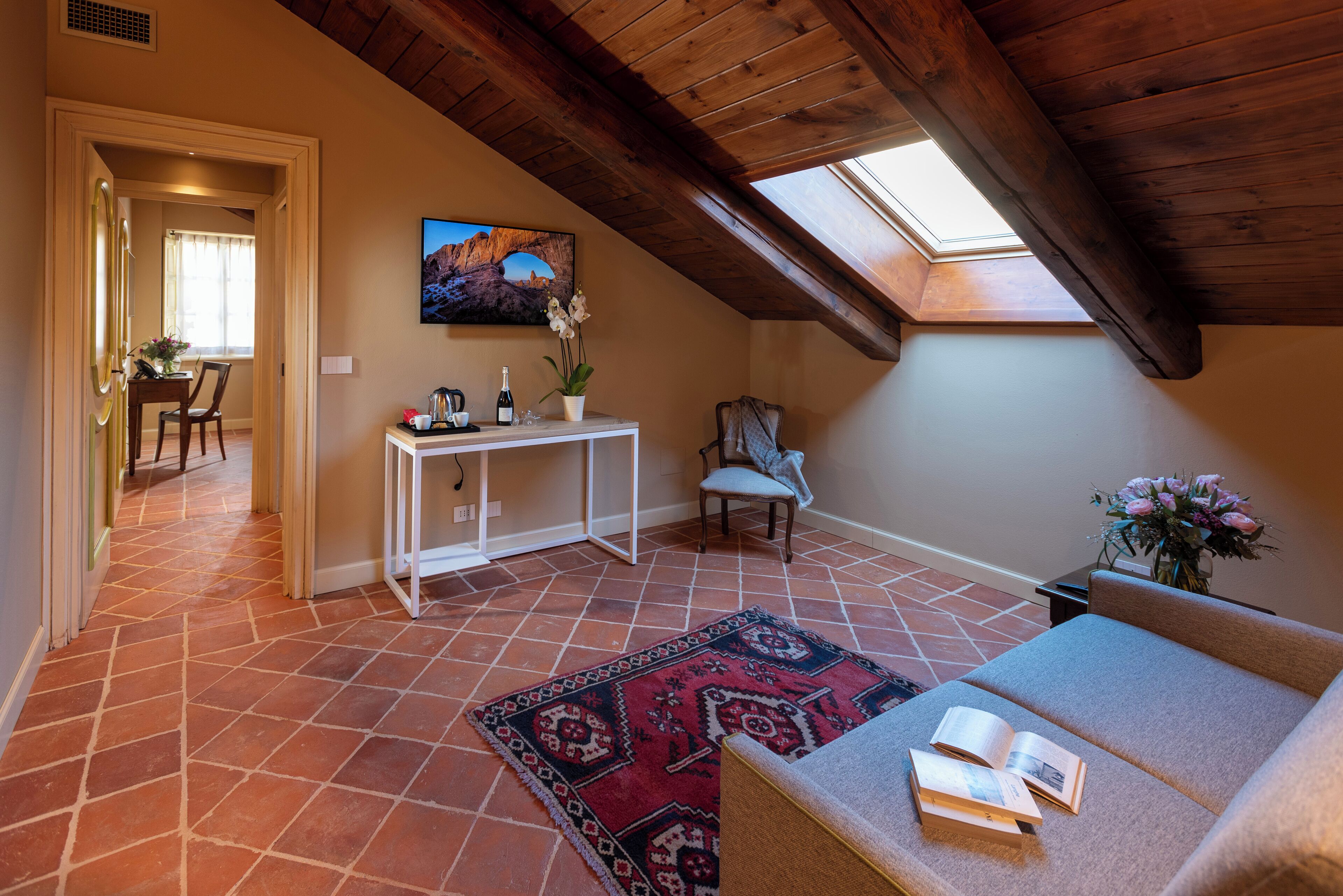 Una camera da letto mansardata accogliente, con soffitto spiovente, un lucernario, un tappeto rosso a motivi e un divano con libri, con un'area pranzo sullo sfondo.