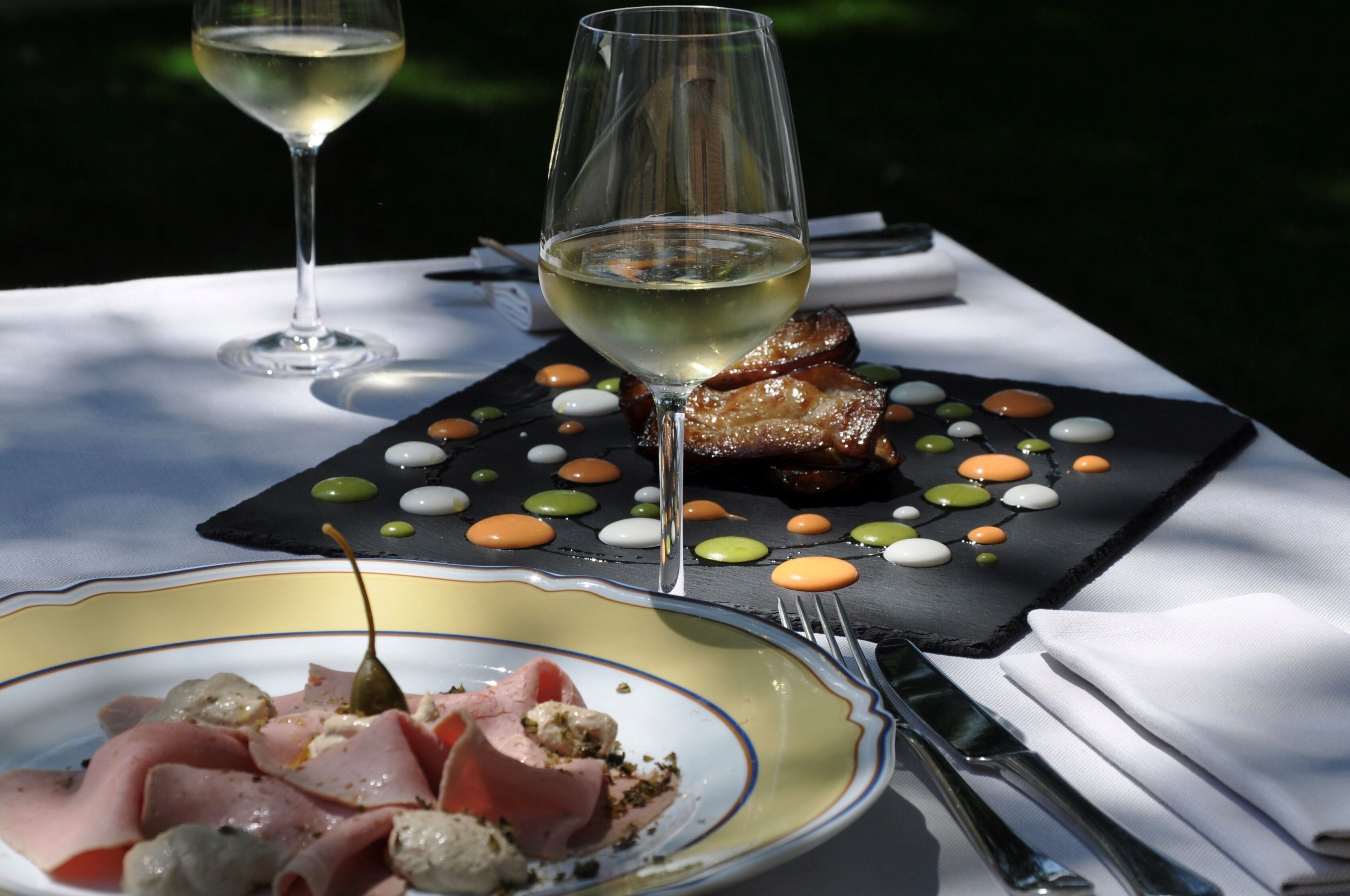 Una mesa de comedor con un plato de comida, una copa de vino blanco y una losa de pizarra negra con puntos de colores.