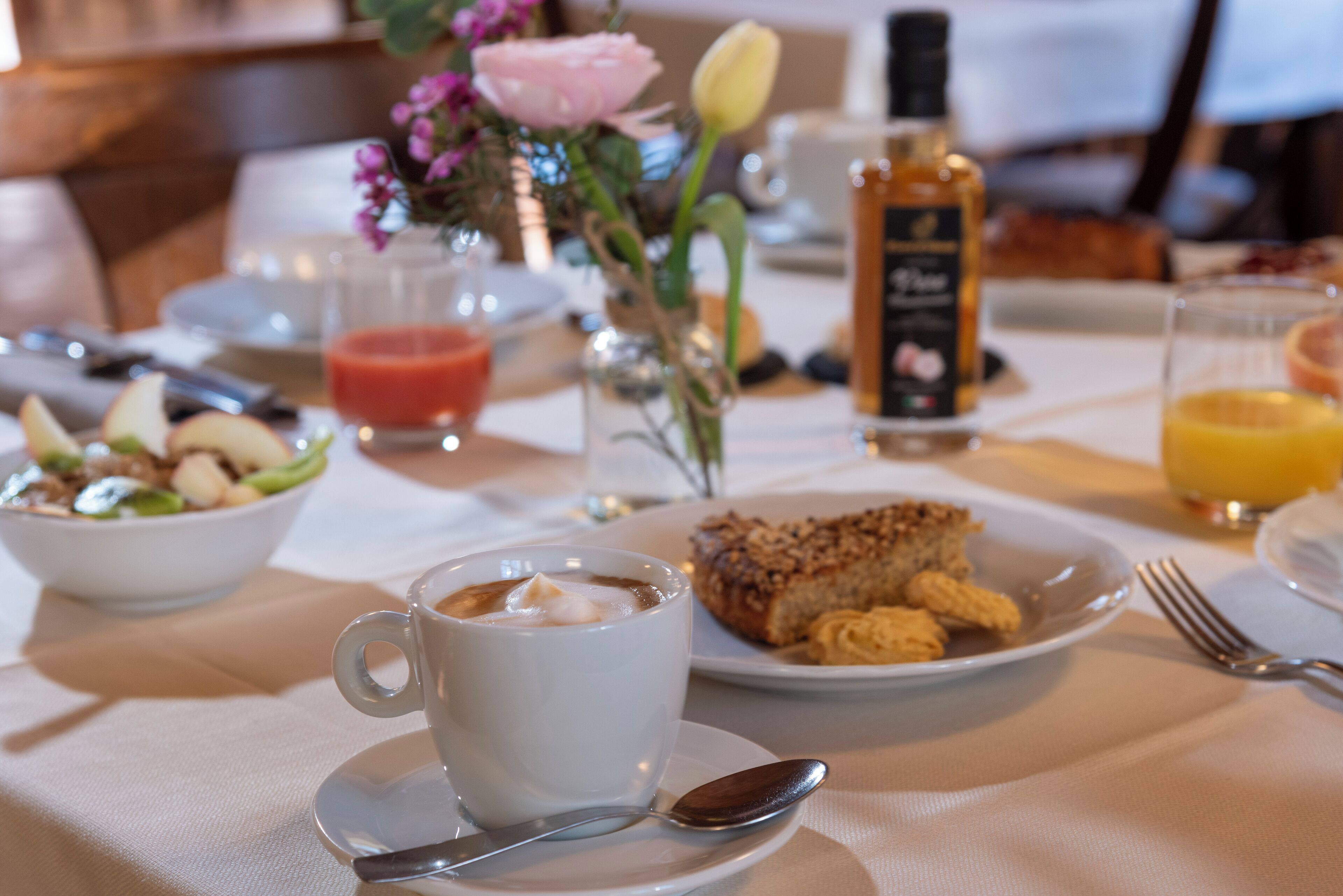 A breakfast setting with a cup of coffee, a plate of pastries, a glass of orange juice, and a floral arrangement.
