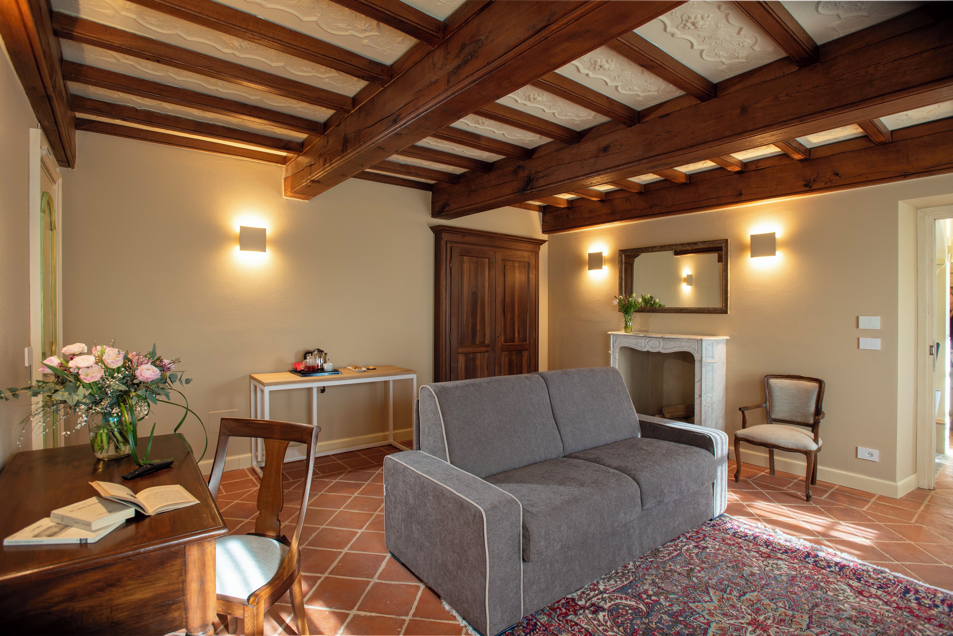 Ein Wohnzimmer mit grauem Sofa, Holzbalkendecke, Kamin und Essbereich mit Tisch und Stühlen.