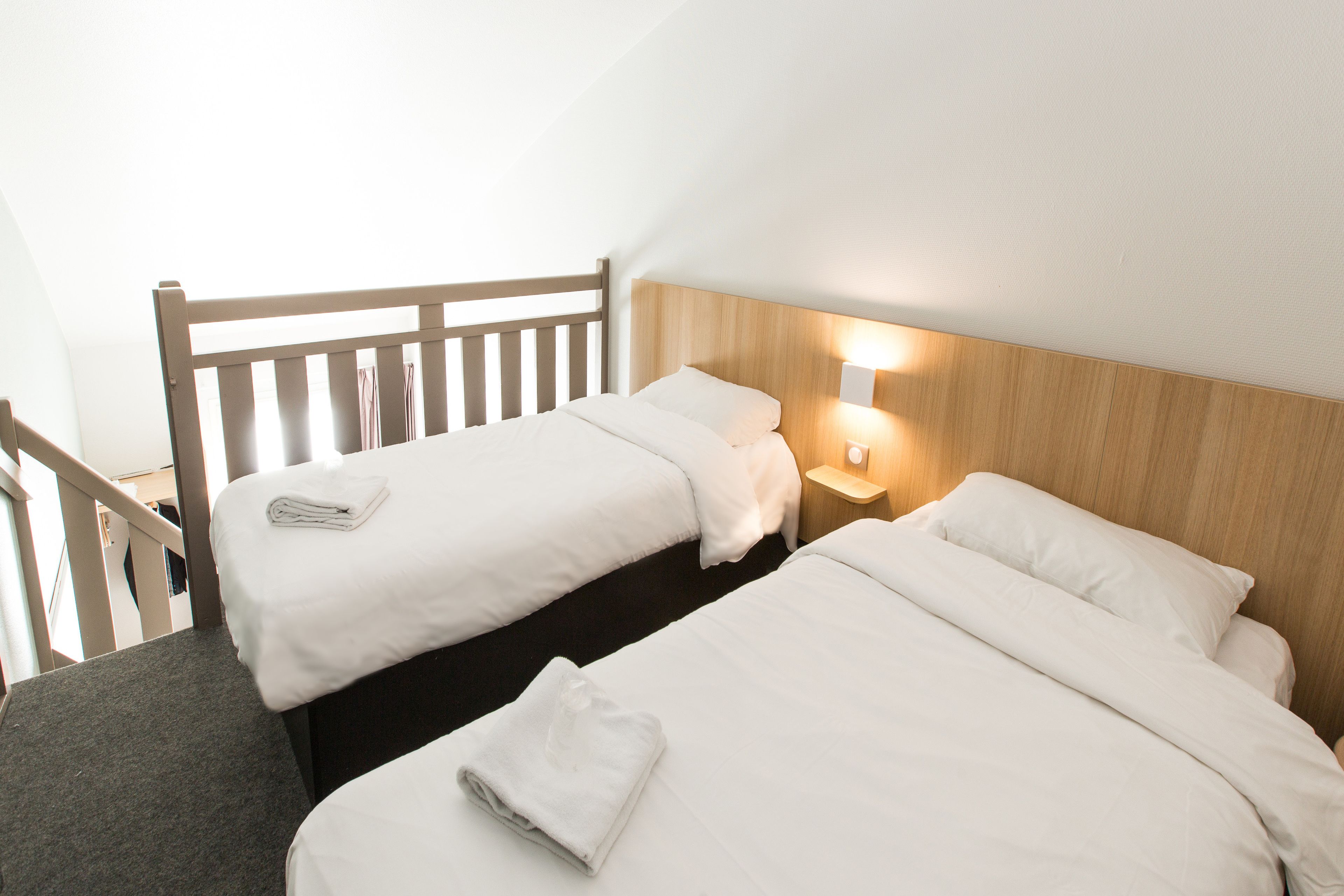 A hotel room with two beds, a wooden headboard, a nightstand with a lamp, and a staircase.