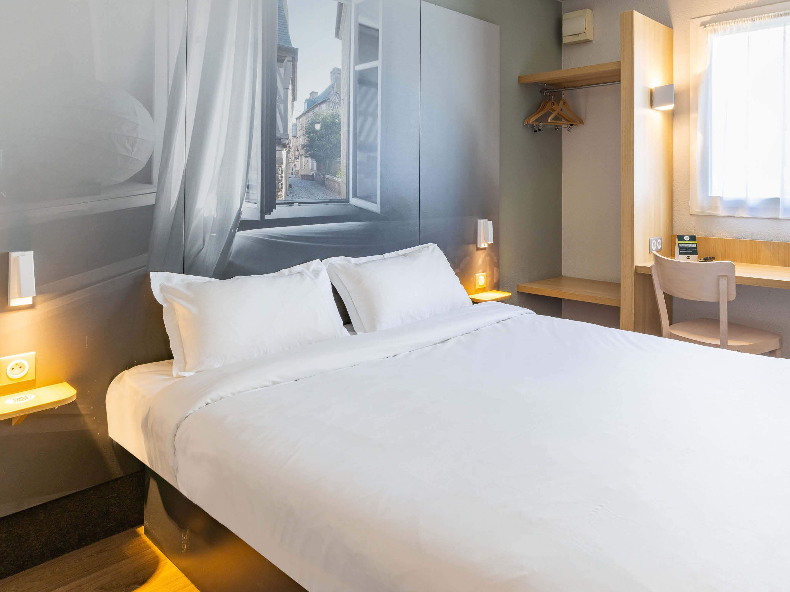 Una cama con sábanas blancas, una mesita de noche con lámpara, una silla de madera y vistas a la calle a través de una ventana.