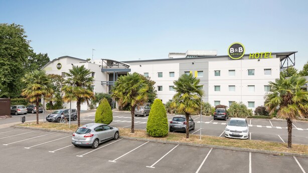 A B&B hotel with a modern design, featuring a parking lot with several cars and palm trees. - B&B HOTEL Rennes Sud Chantepie (Rennes)