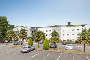 A B&B hotel with a modern design, featuring a parking lot with several cars and palm trees. - B&B HOTEL Rennes Sud Chantepie (Rennes)