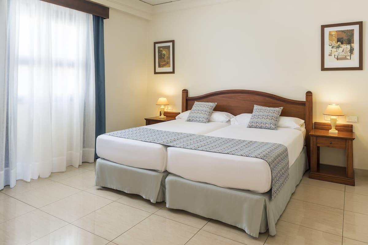 A double bed with two pillows, a bedside table with a lamp, and a window with curtains.