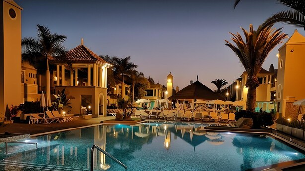 Une piscine entourée de palmiers et de bâtiments dont l’éclairage chaleureux se reflète au crépuscule, avec des chaises longues et des parasols à proximité.