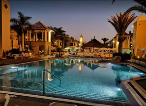 Ein Swimmingpool mit Liegestühlen und Sonnenschirmen, umgeben von Palmen und Gebäuden mit warmem Schein im Abendlicht.