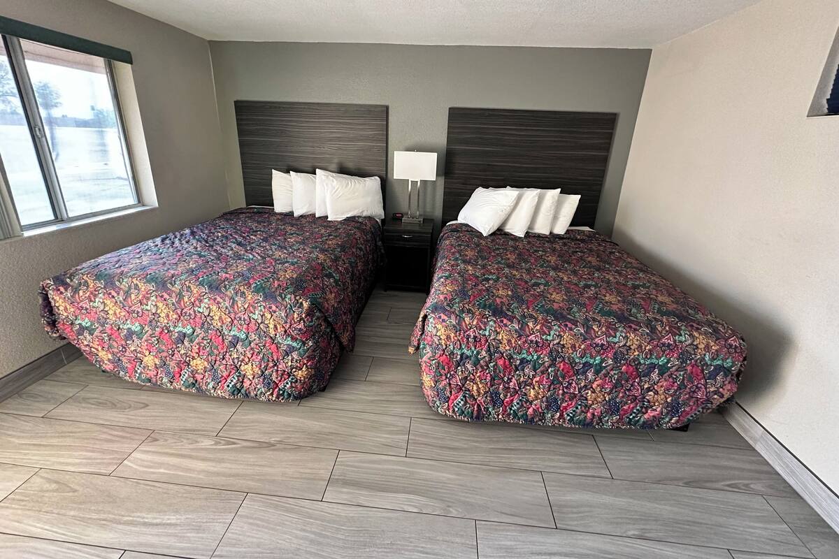 A hotel room with two beds, a nightstand with a lamp, and a window with a view of the outside.
