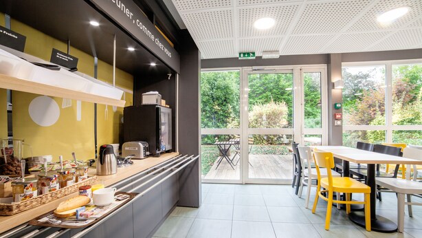 Een ontbijtruimte met een toonbank waarop etenswaren staan, een koffieapparaat en een zithoek met tafels en stoelen.