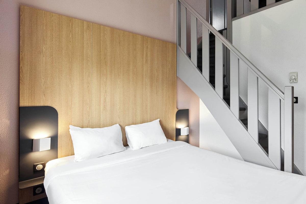 Una habitación de hotel moderna con un cabecero de madera, una cama con ropa blanca, dos lámparas de noche y una escalera con barandilla de cristal.