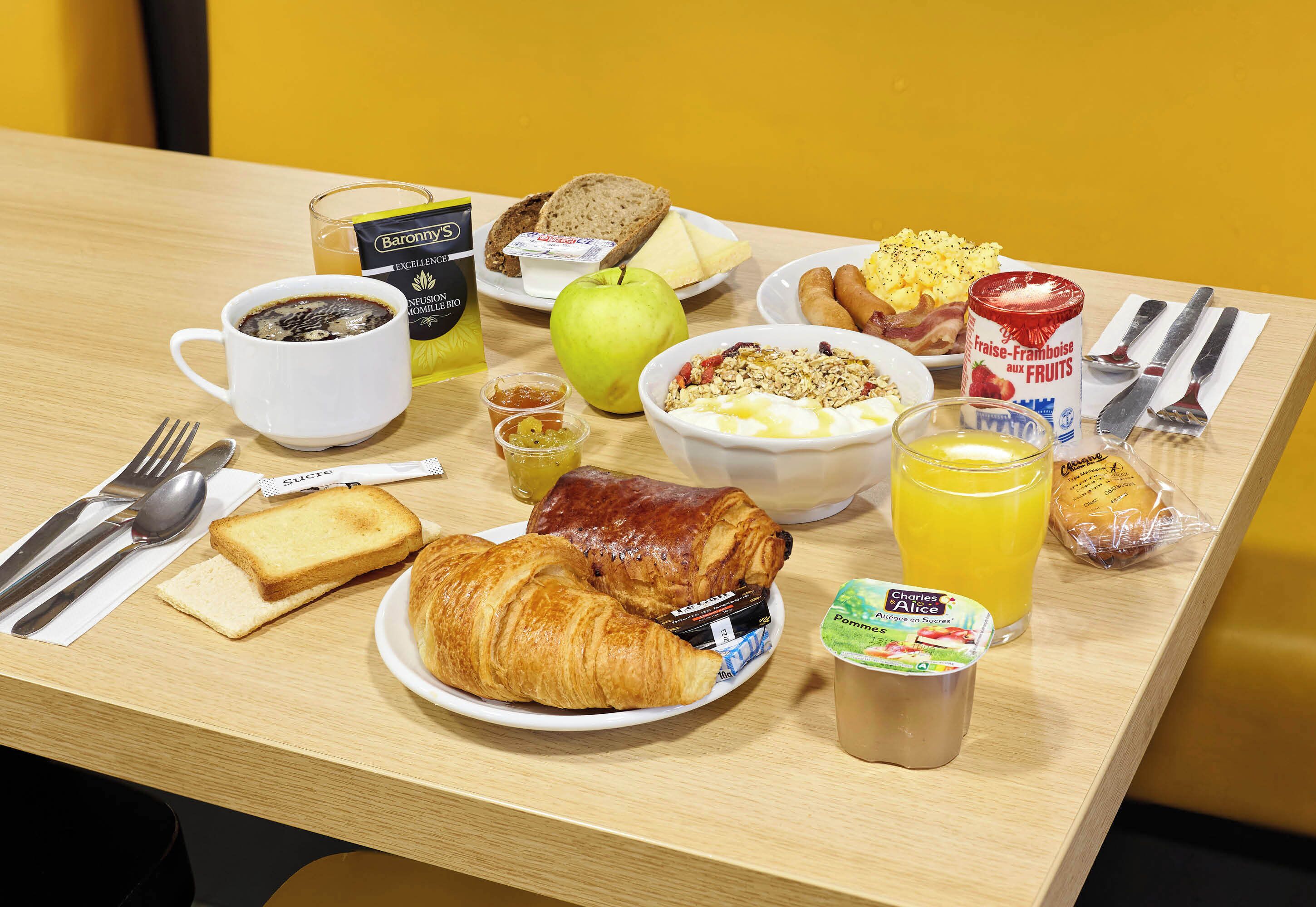 Ein Frühstückstisch mit Croissants, Toast, einem Croissant-Sandwich, einer Schüssel mit Müsli, einem Glas Orangensaft, einer Tasse Kaffee, einer Dose Früchte und einer Milchflasche.