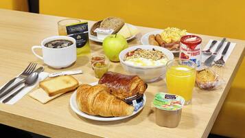 Ein Frühstückstisch mit Croissants, Toast, einem Croissant-Sandwich, einer Schüssel mit Müsli, einem Glas Orangensaft, einer Tasse Kaffee, einer Dose Früchte und einer Milchflasche.