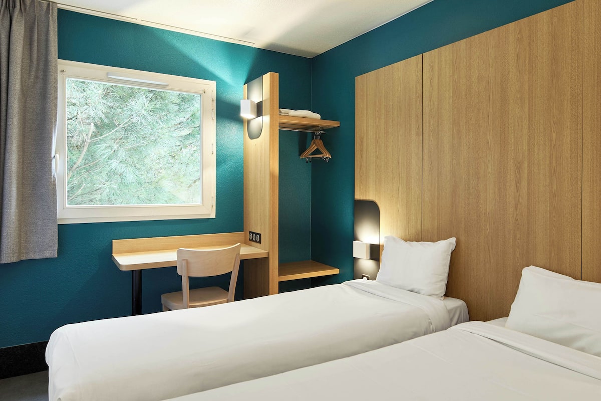 Habitación de hotel con dos camas, un escritorio, una silla, una ventana con vistas a árboles y un armario de madera.