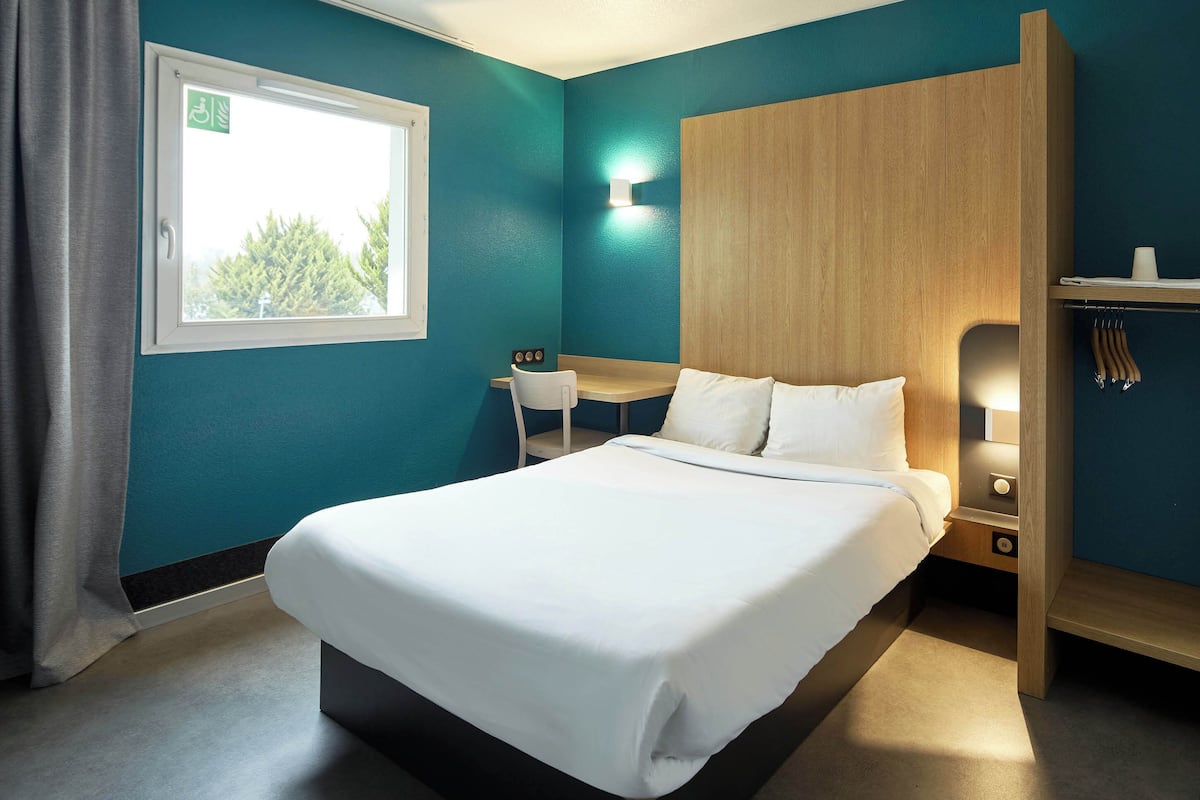 Habitación de hotel con cama, escritorio, silla, armario y ventana con vista a árboles.
