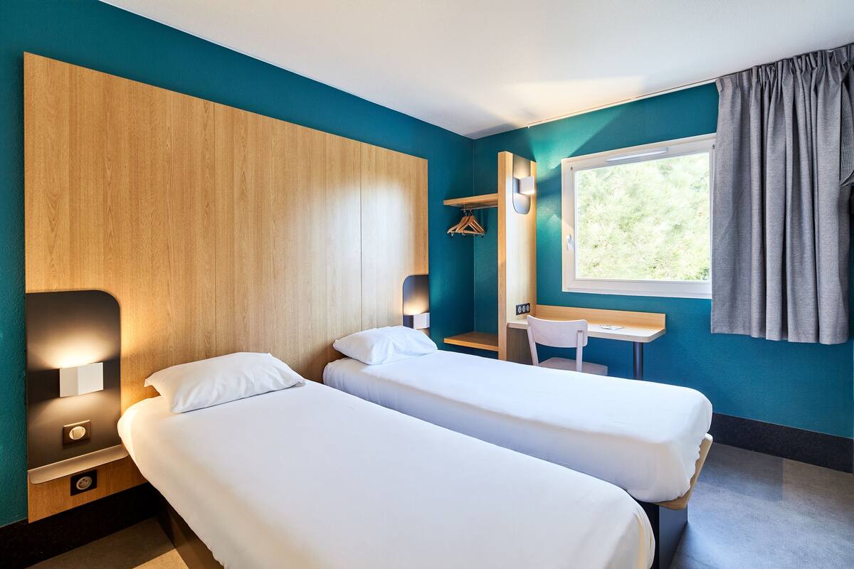 Habitación de hotel con dos camas, un escritorio, una silla, un ventanal con cortinas y cabecero de madera.