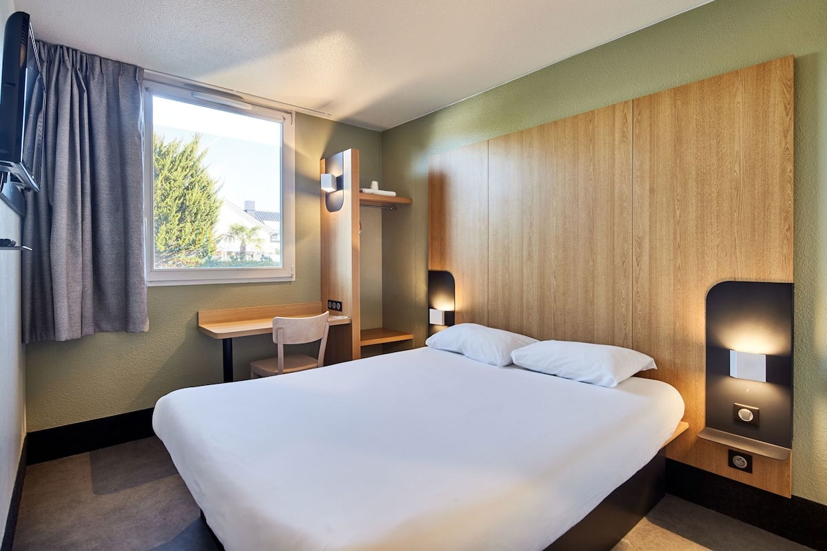 Habitación de hotel con una cama grande, un escritorio, una silla, un armario y una ventana con cortinas.