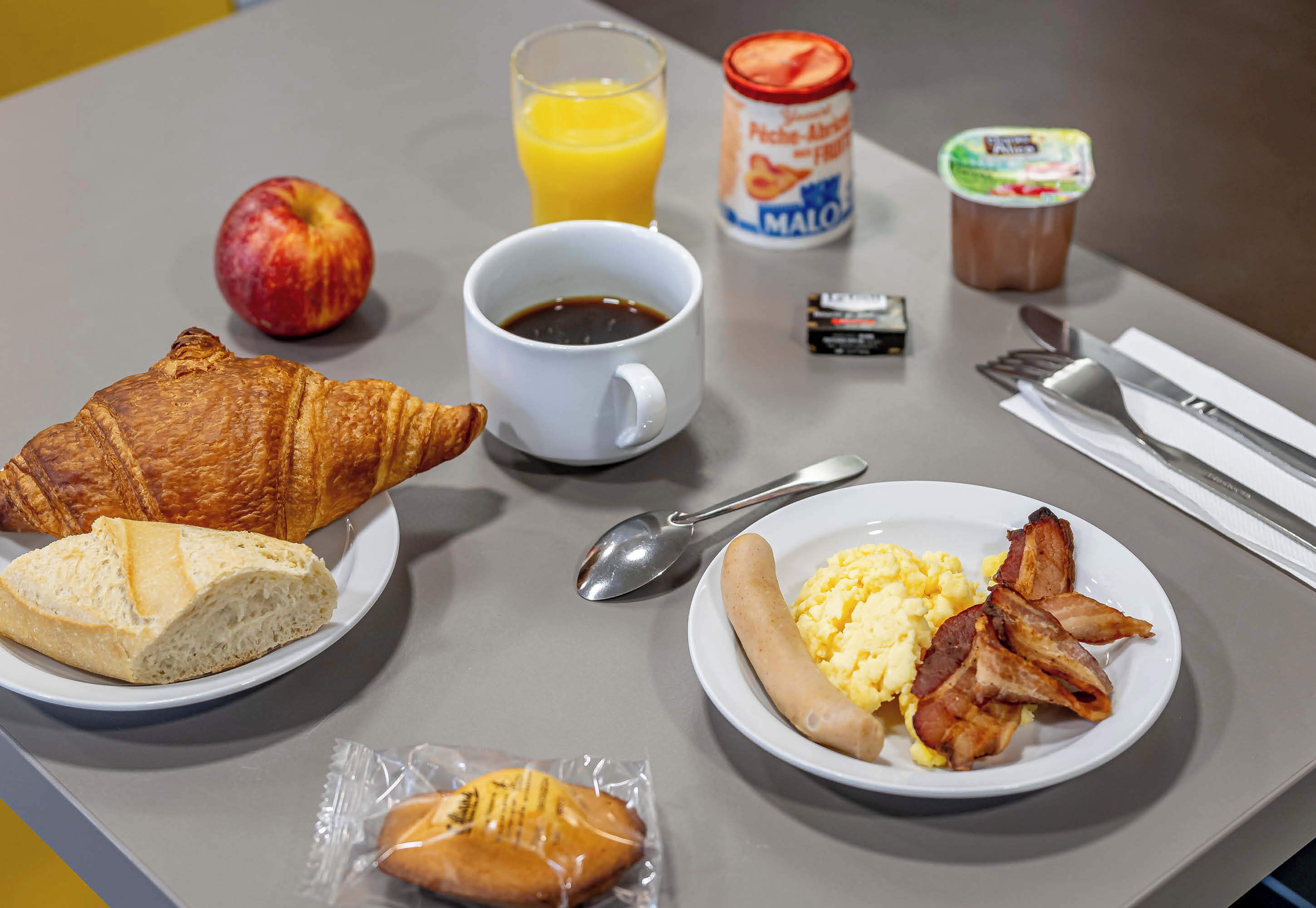 Una tavola imbandita per la colazione, con croissant, una porzione di bacon e uova, un bicchiere di succo d'arancia, una lattina di Mambo, un piccolo pacchetto di burro e una tazza di caffè.